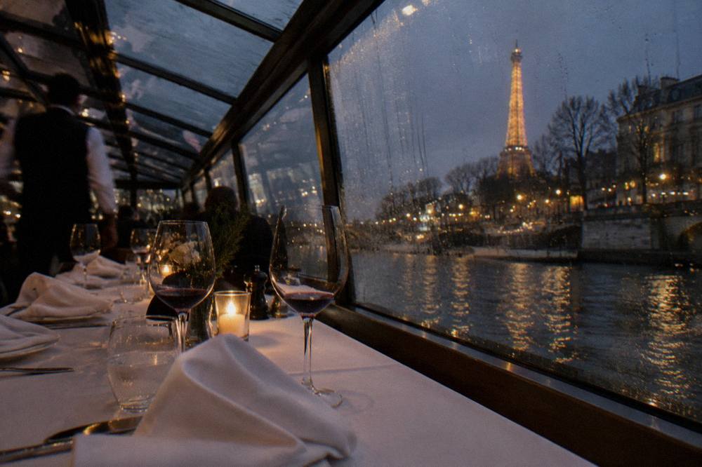 A set dinner table with wine glasses inside a glass-roofed boat cabin, city lights visible through the glass