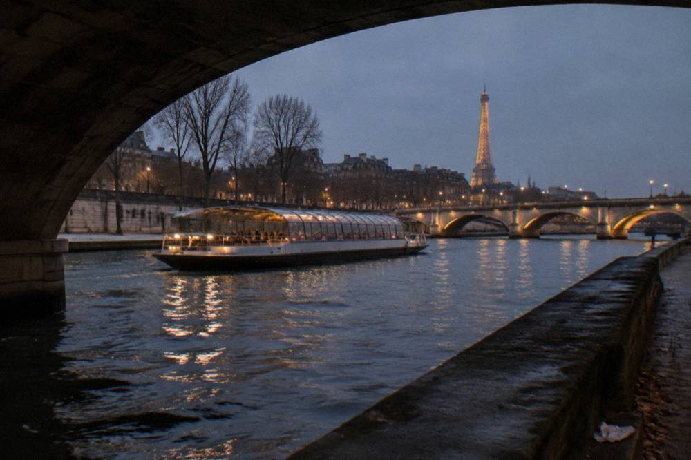 A covered dinner boat passing illuminated bridges on the Seine at dusk in December.