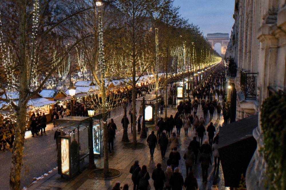 An illuminated city avenue at night with trees wrapped in white lights and a dense evening crowd walking below