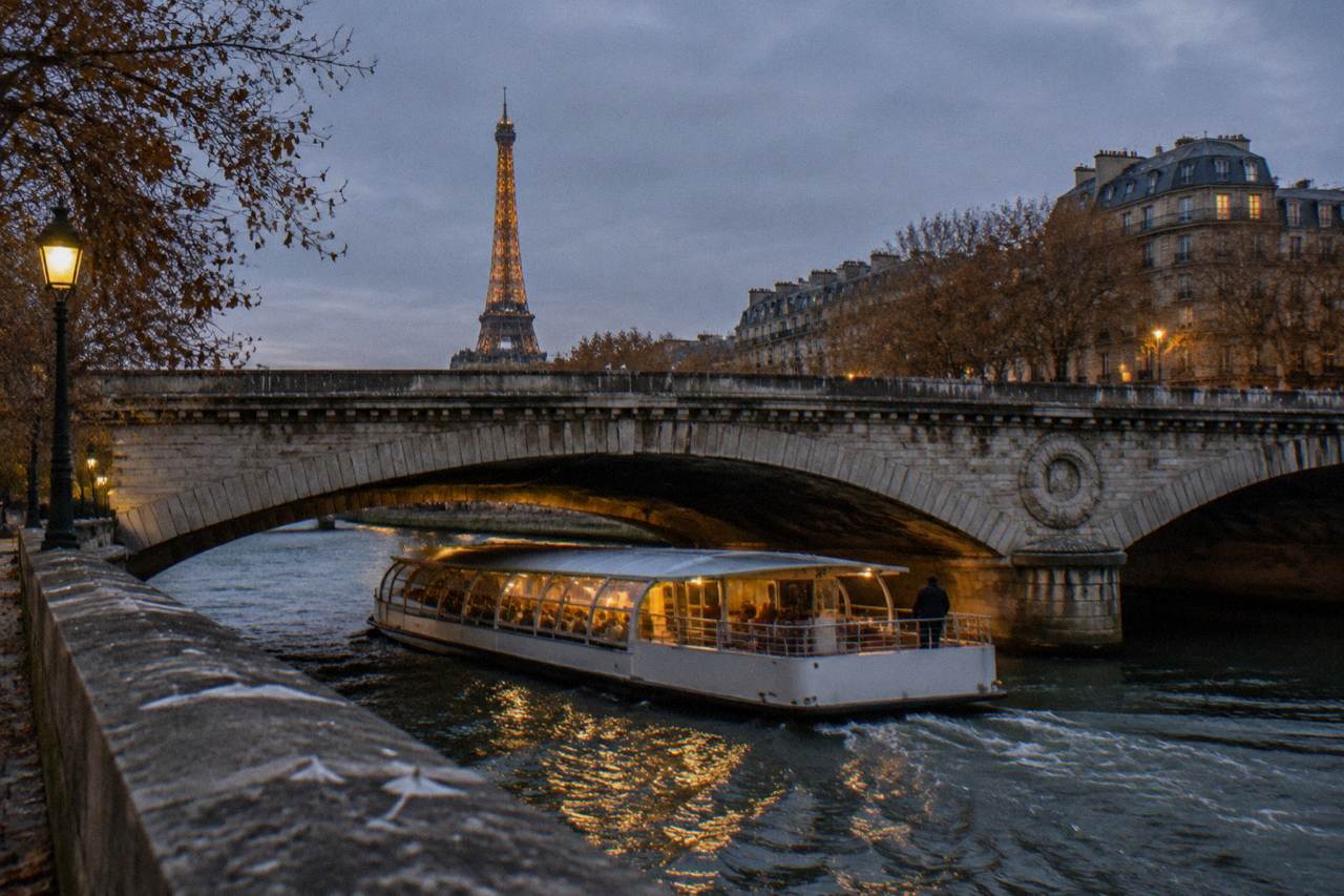 A covered passenger boat passing under a stone bridge on the Seine at dusk