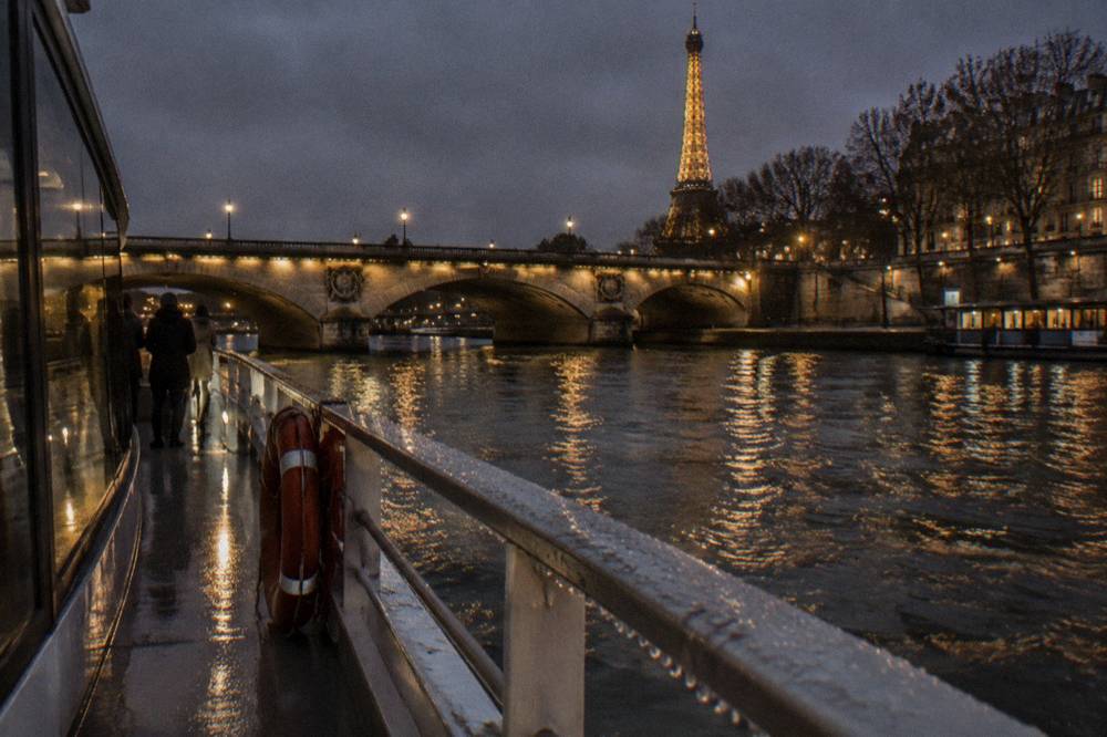 A lit bridge over the Seine seen from a boat deck in winter, wet railings and city glow