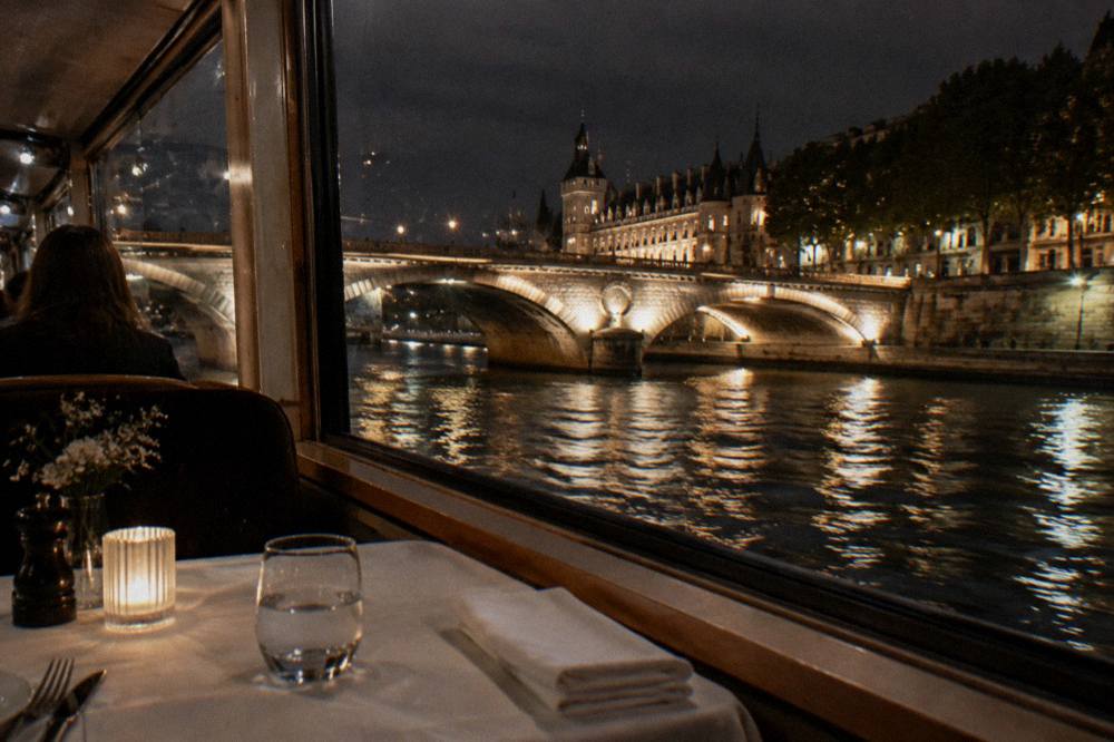 A dinner table set beside a window overlooking the illuminated Seine at night