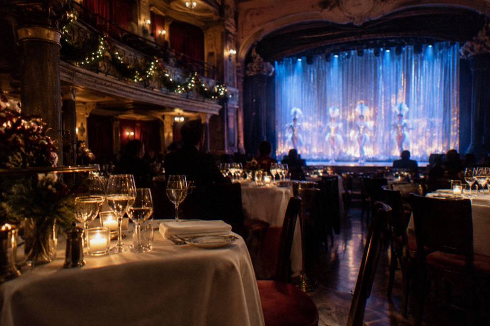 Dinner tables set with glassware in a cabaret theatre facing an illuminated stage