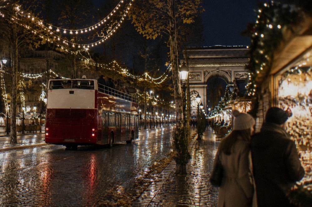 An open-top sightseeing bus passing under lit Christmas garlands on a Paris boulevard
