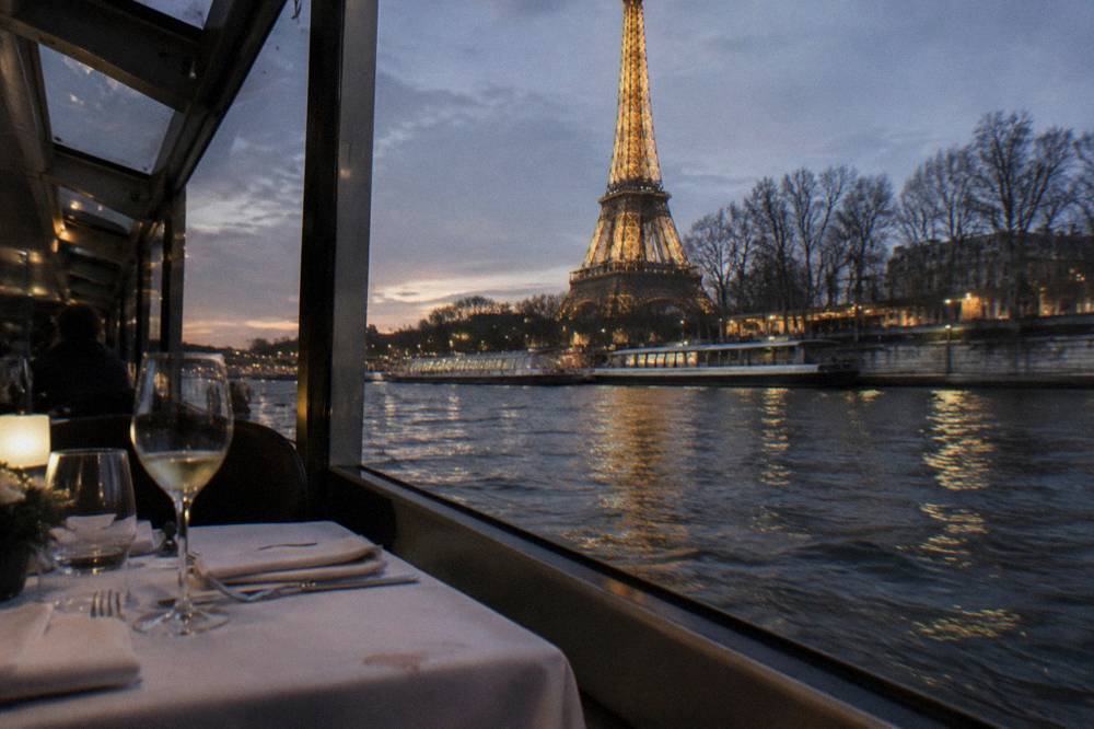 A set dinner table with glasses and folded napkins beside a window overlooking a dark river