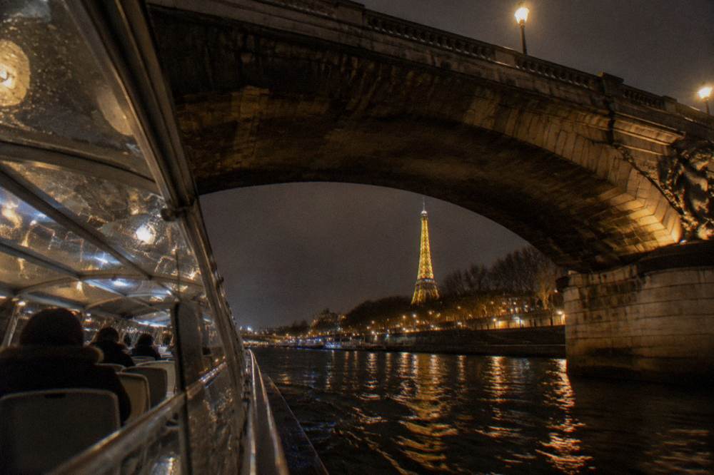 Cruise boat passing under an illuminated Paris bridge at night with lights reflecting on the Seine