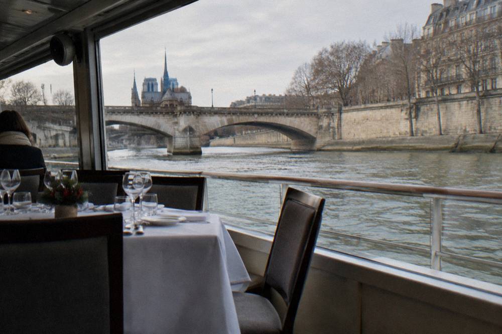 Lunch table set on a covered boat with the Seine and a stone bridge visible through the windows