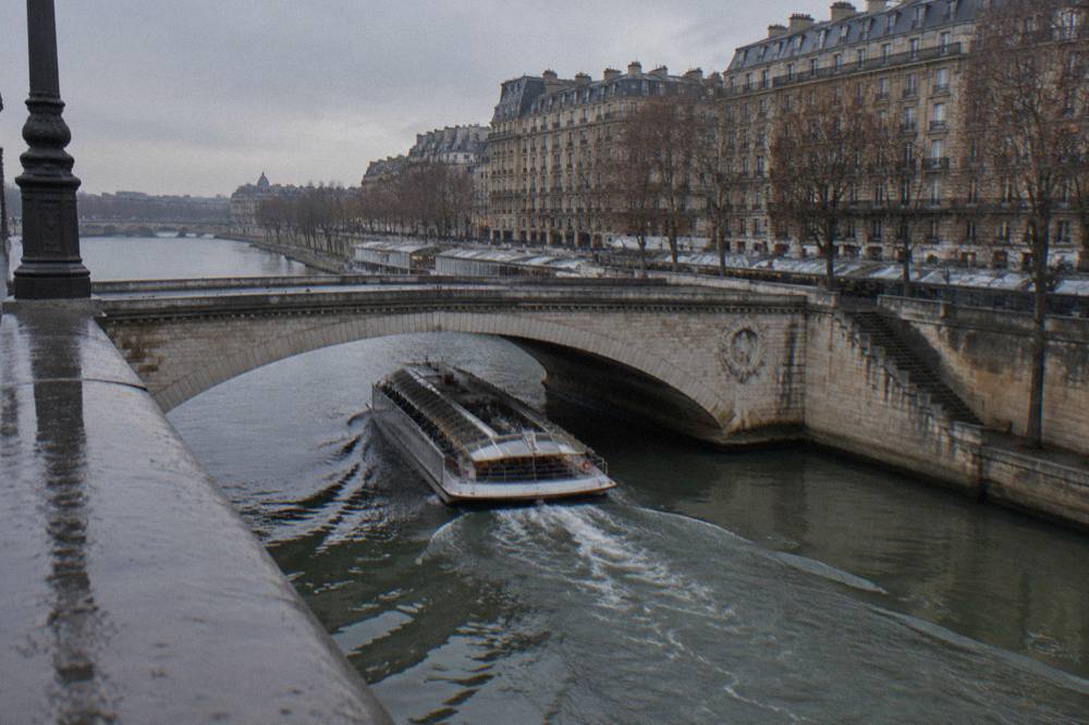 A glass-canopy river boat on the Seine at dusk with illuminated bridge arches and city lights reflected in the water