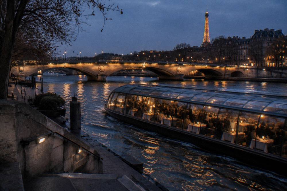 A glass-roofed dinner boat on the Seine passing illuminated bridges at night
